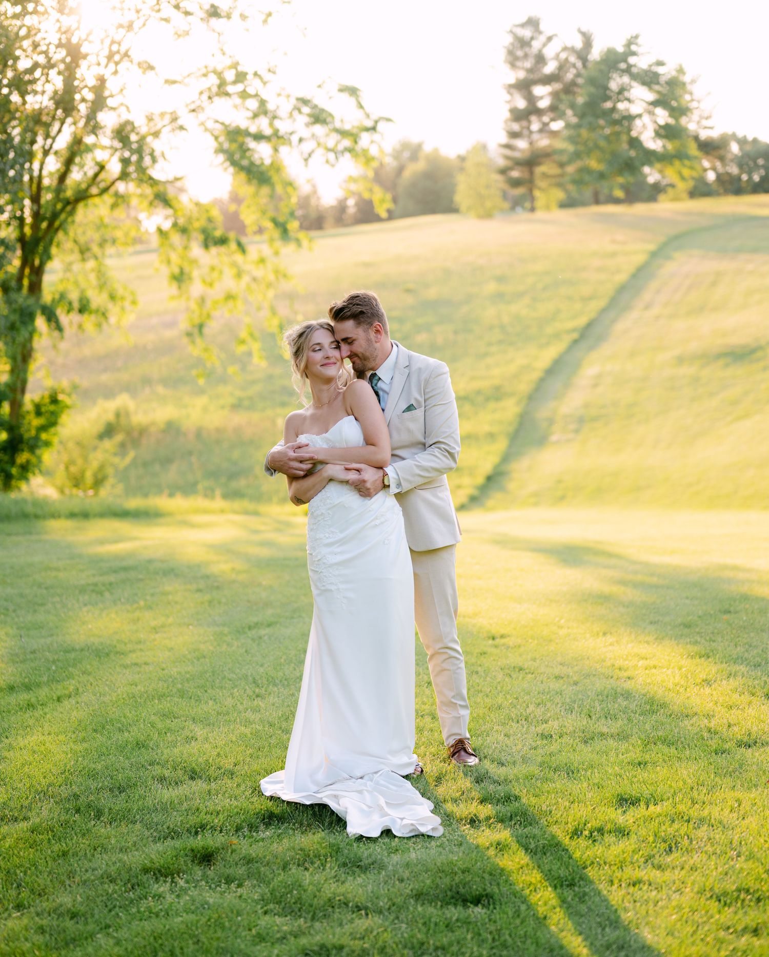 Bride and groom embrace on lush golf course lawn during golden hour sunset wedding portrait.