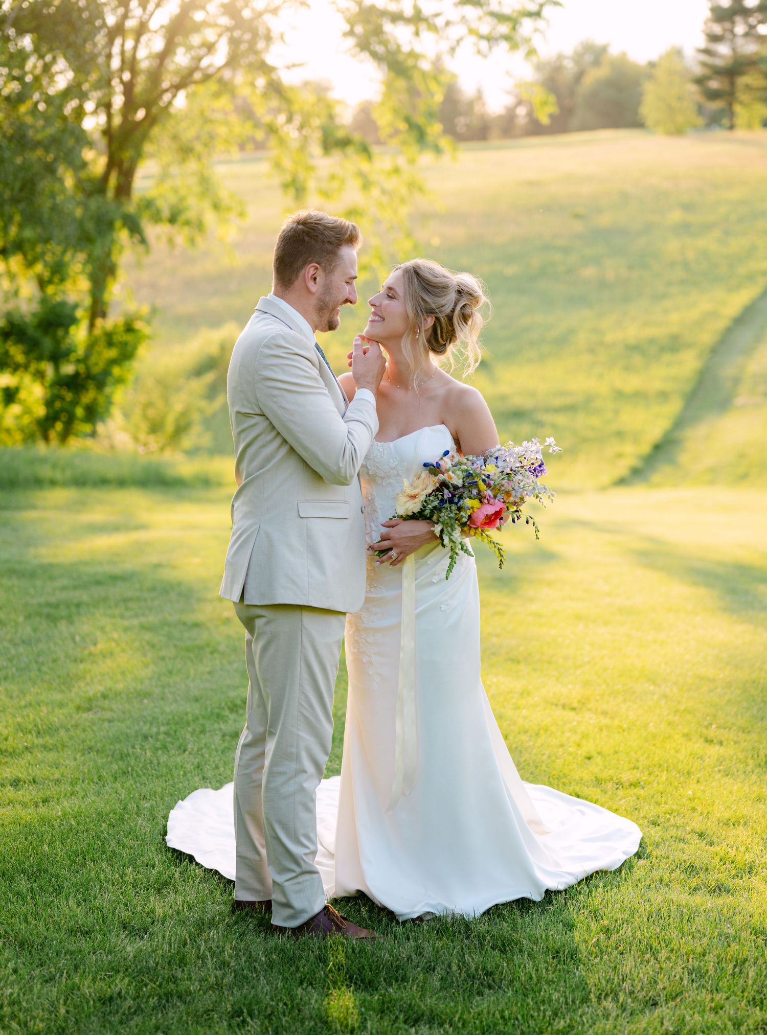 Bride and groom share romantic golden hour moment on green hillside, bride holds colorful bouquet.