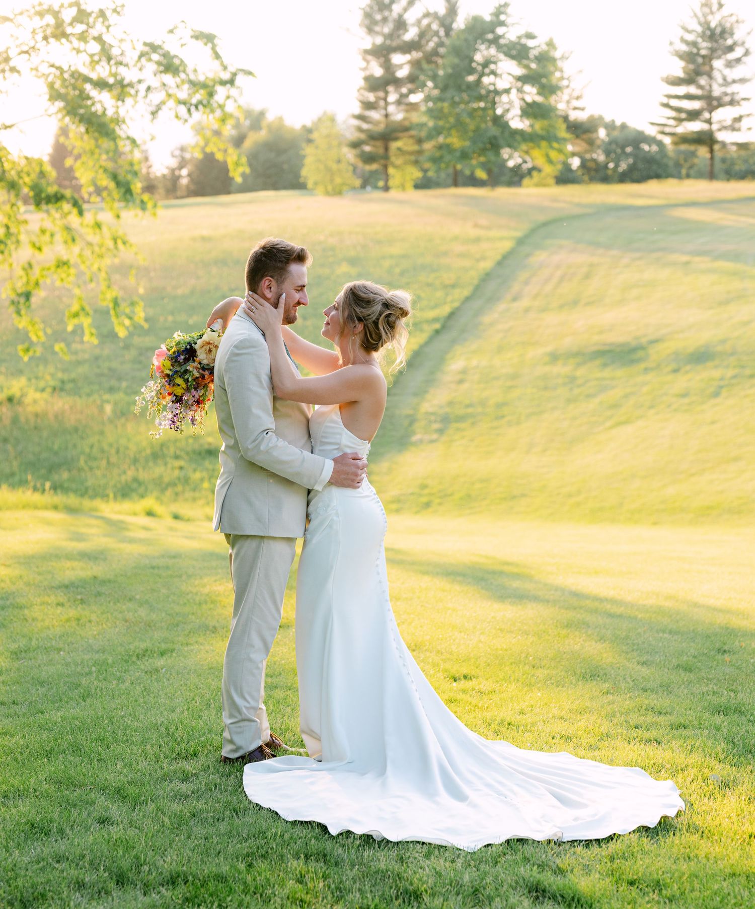 Newlyweds embrace on golf course at sunset, bride in off-shoulder gown holding vibrant bouquet.