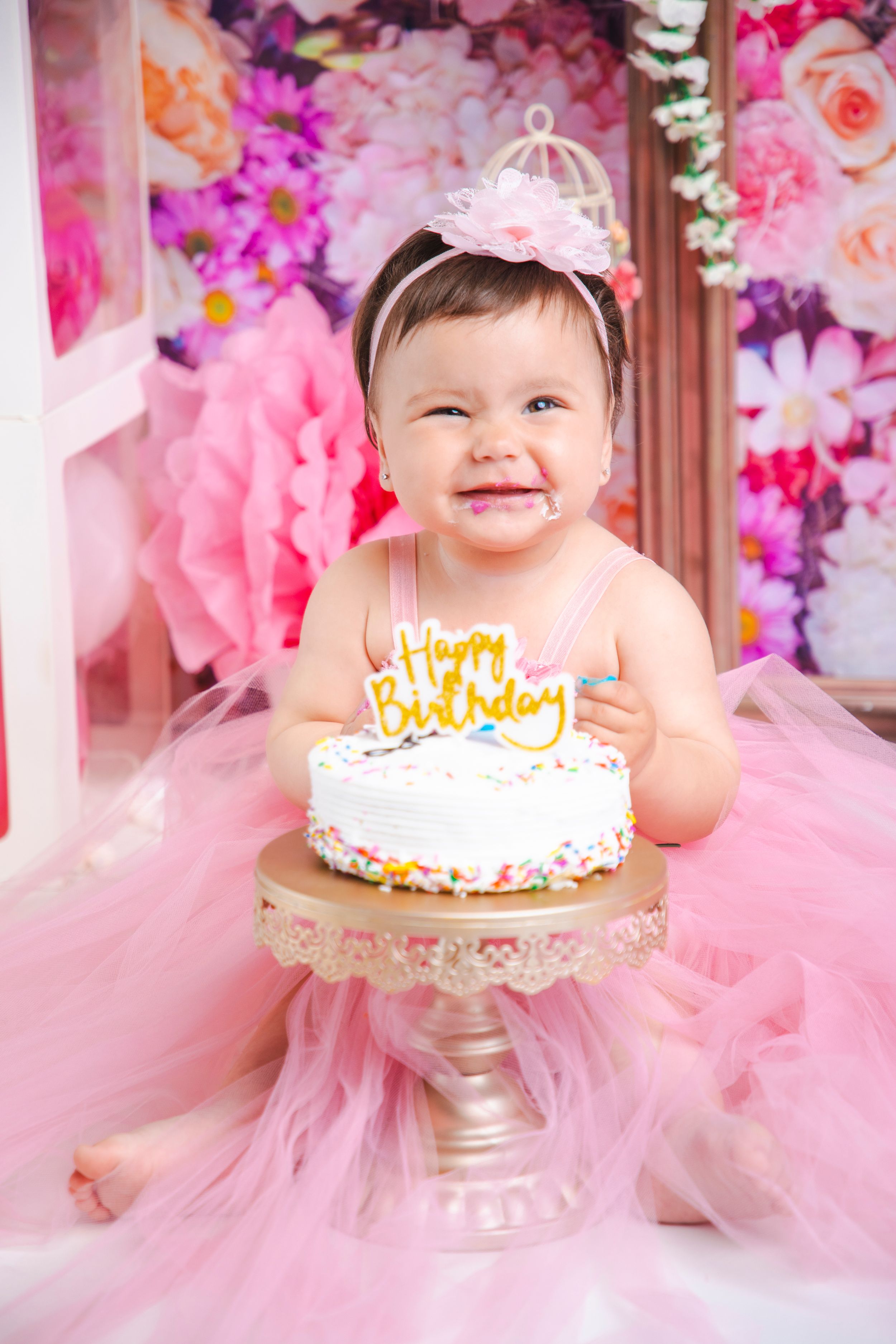 Birthday celebration setup with cake on pedestal against pink floral backdrop.