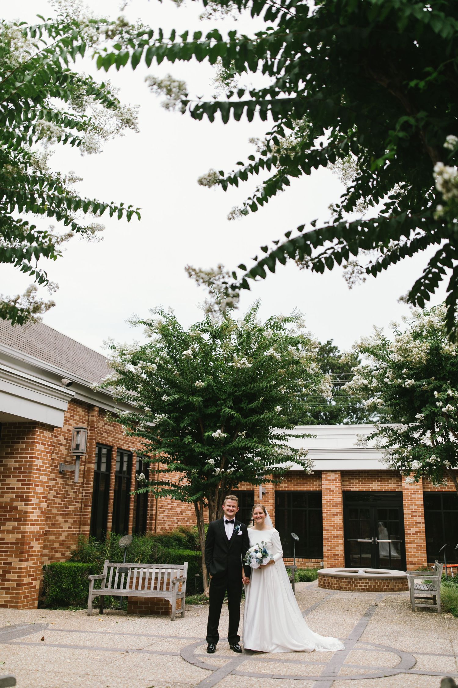 A wedding couple poses for a portrait in front of a brick building surrounded by leafy trees.