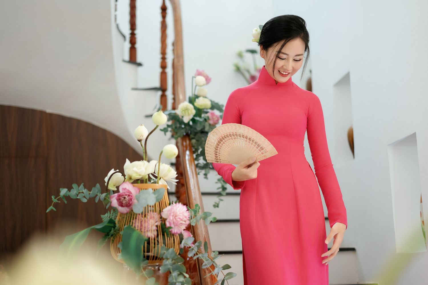 Woman in red traditional dress holding fan amidst floral decorations on staircase.