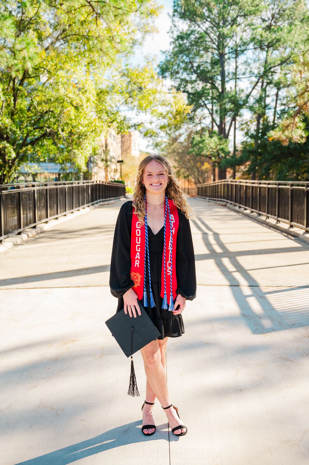 Columbus State University Graduate wearing red Cougar Athletics graduation stoles with a black v-neck dress poses outdoors.