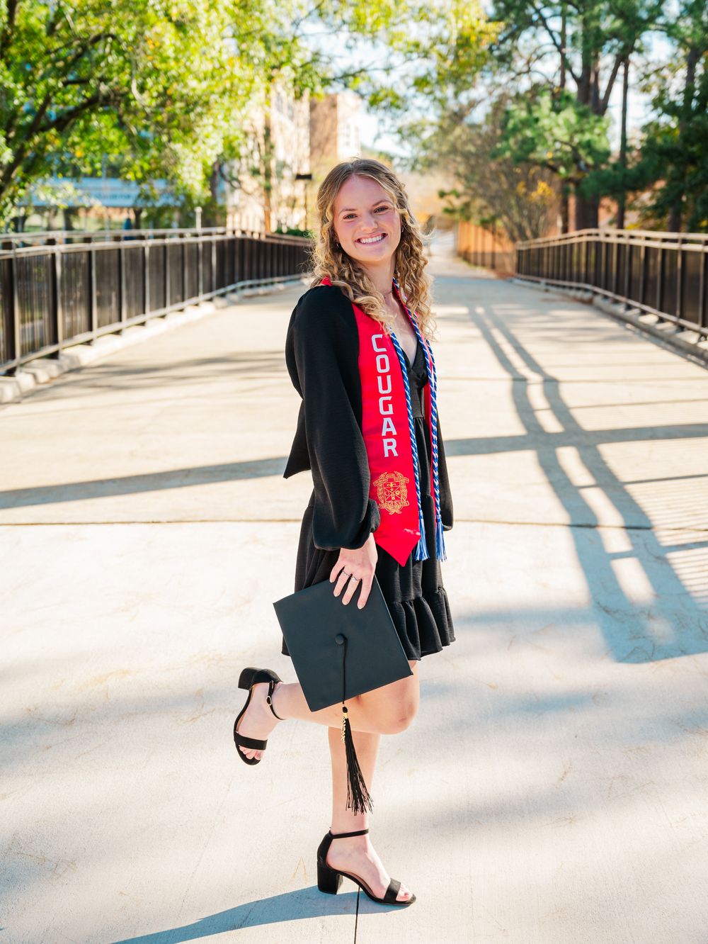 Columbus State University Graduate wearing red Cougar Athletics graduation stoles with a black v-neck dress poses outdoors.