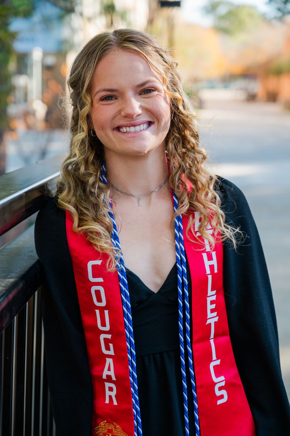 Columbus State University Graduate wearing red Cougar Athletics graduation stoles with a black v-neck dress poses outdoors.