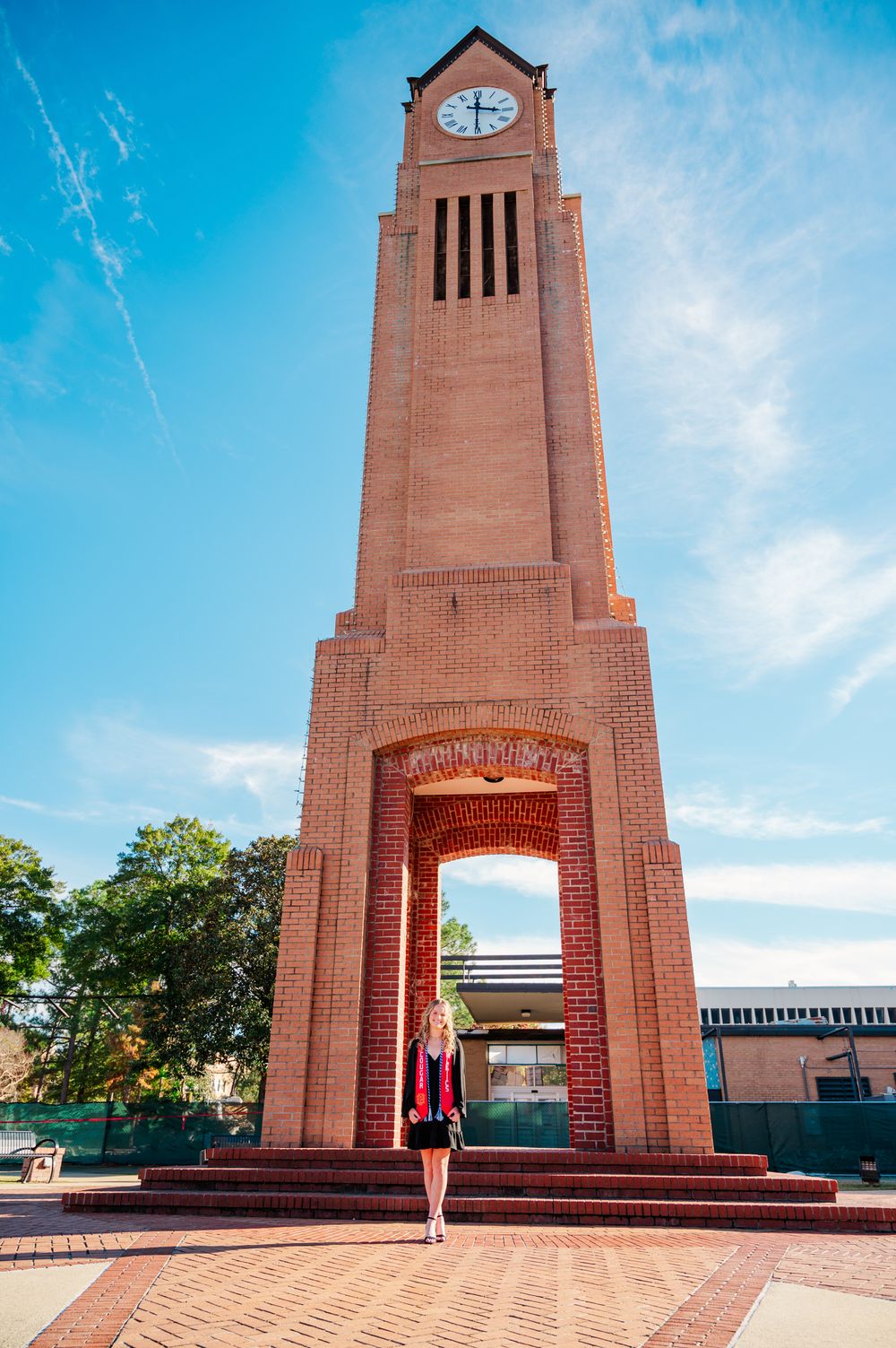 CSU Graduate in red and black regalia stands under a brick archway on a sunny day at university campus.