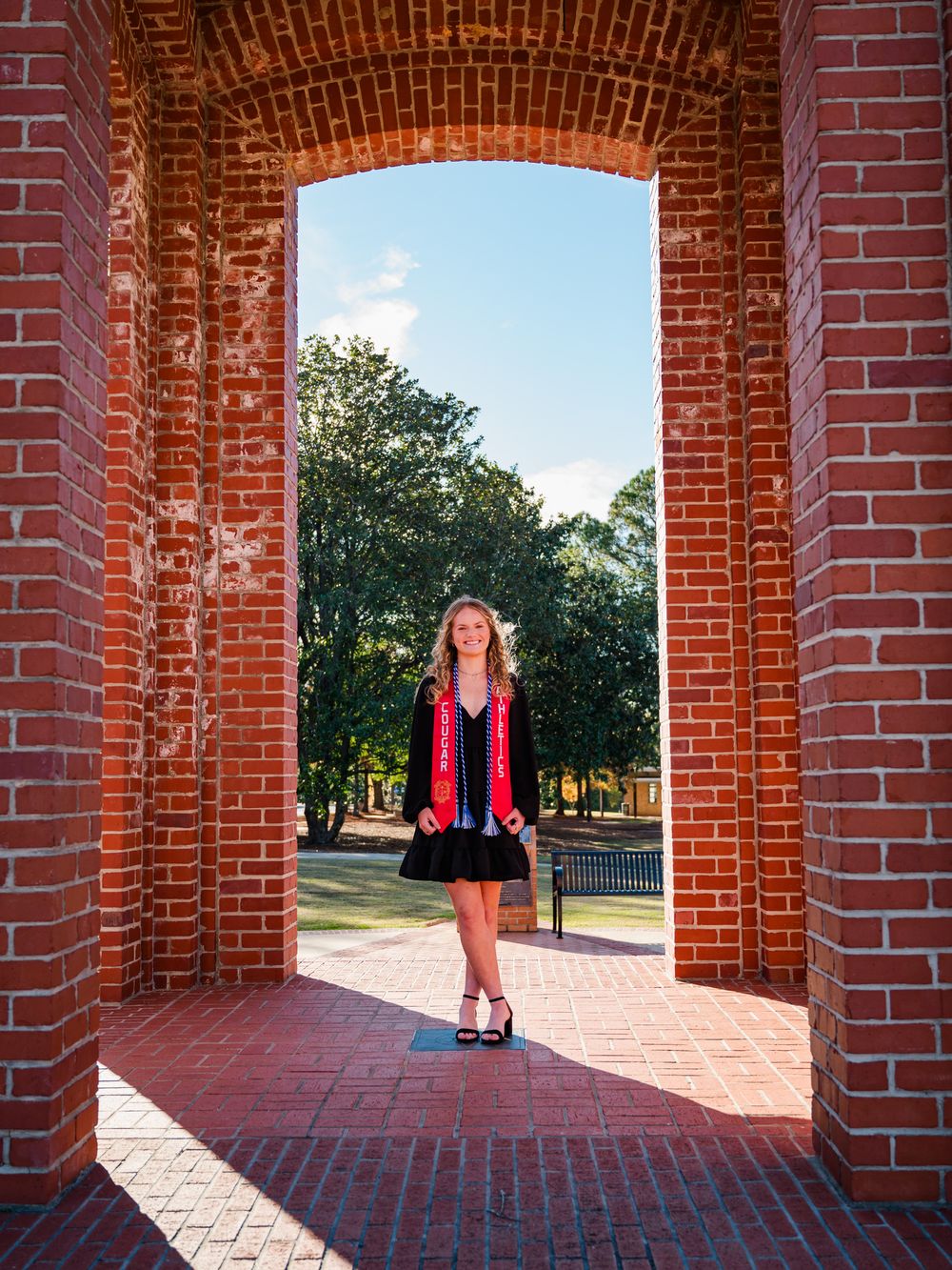 Graduate in red and black regalia stands under a brick archway on a sunny day at university campus.