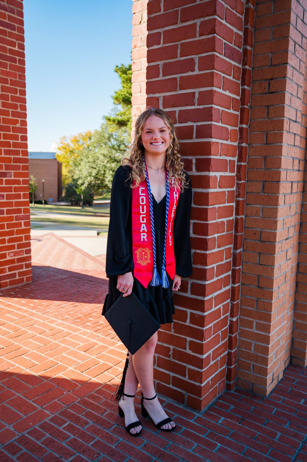 Graduate in black academic gown and red honors sash stands between brick columns on a sunny day.
