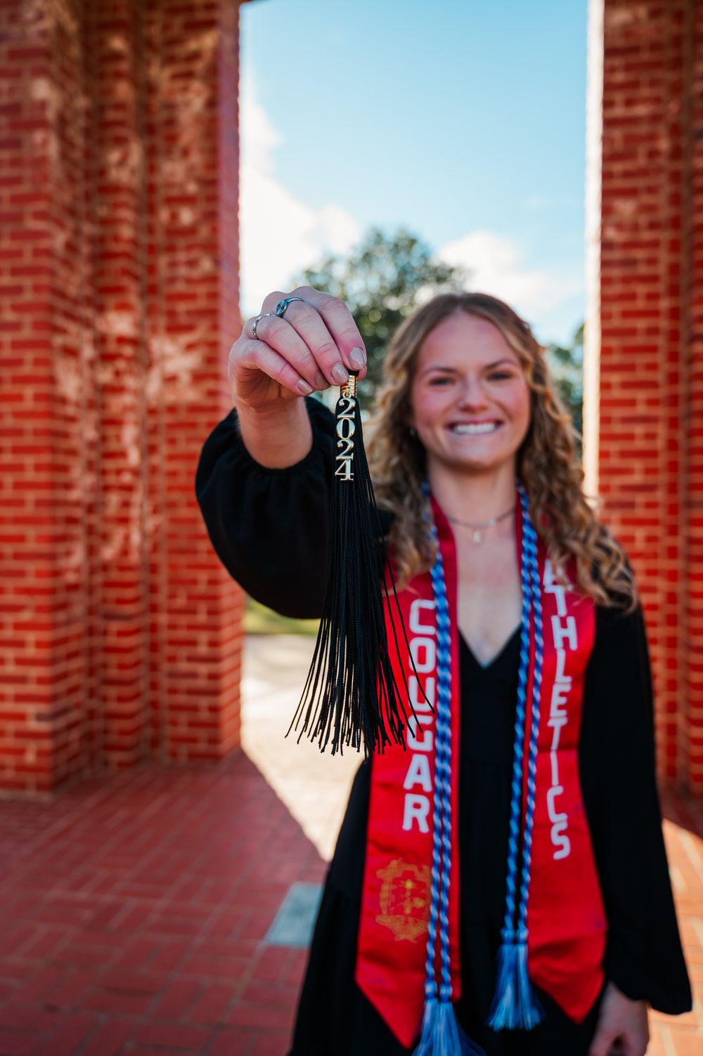 CSU Graduate in cap and gown holds up graduation tassel with 2024 charm while wearing honor cords and red stole outdoors.