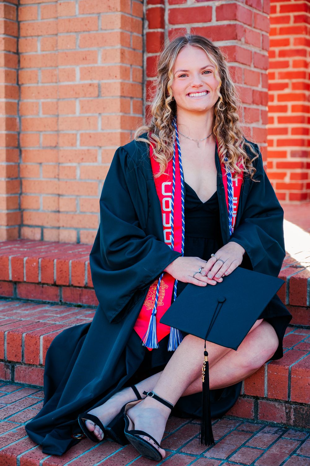 Columbus State University Graduate in black cap and gown with red stole sits smiling on brick steps during commencement photo session.