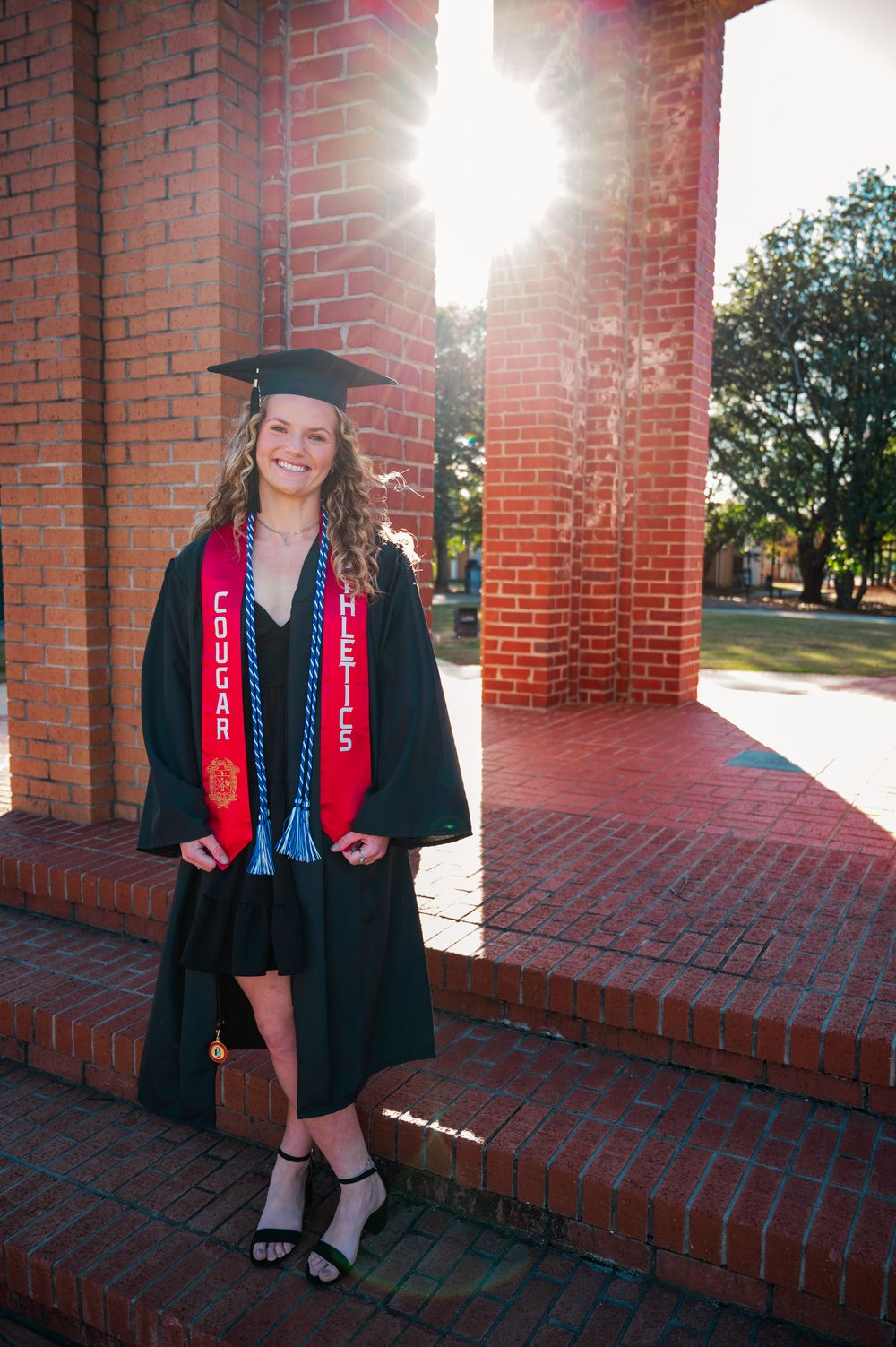 A Columbus State University graduate in cap and gown with honor cords poses by a brick building with sunlight streaming down.