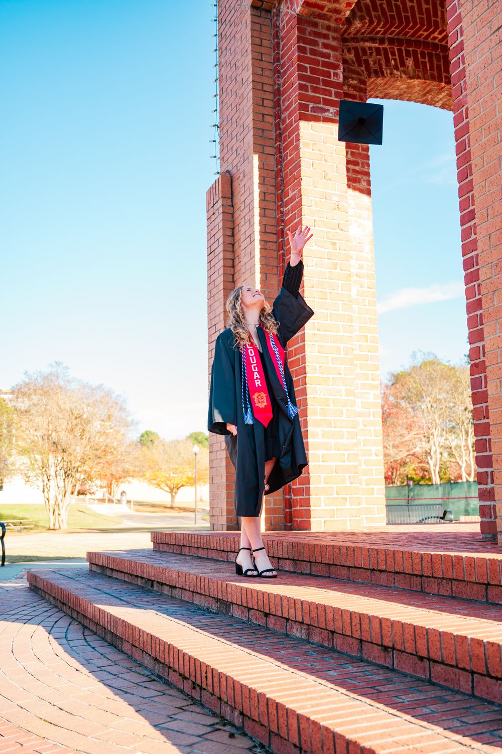 Columbus State University Graduate wearing traditional black academic gown poses by brick column outdoors on sunny day holding diploma.