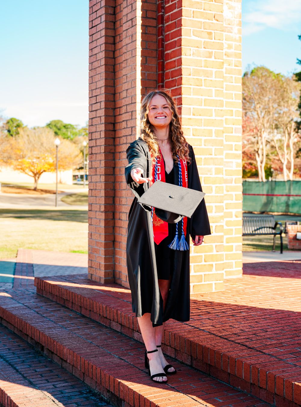 Columbus State University Graduate wearing traditional black academic gown poses by brick column outdoors on sunny day holding diploma.
