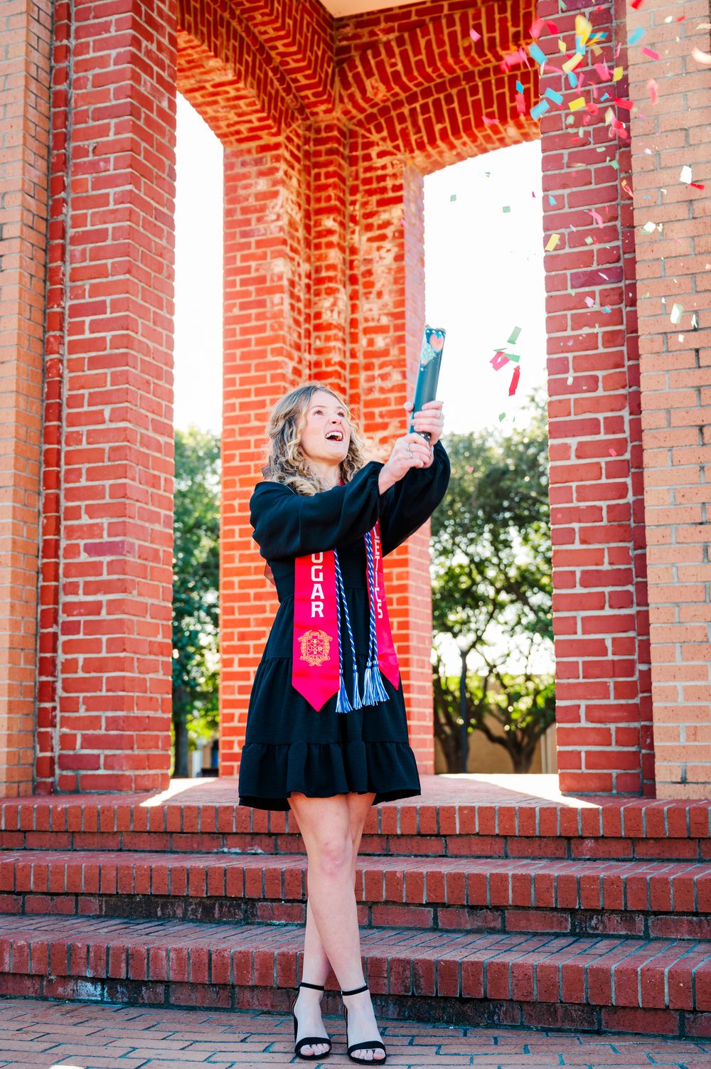 A CSU graduate in a black dress with red sash celebrates on brick steps while tossing confetti in the air.