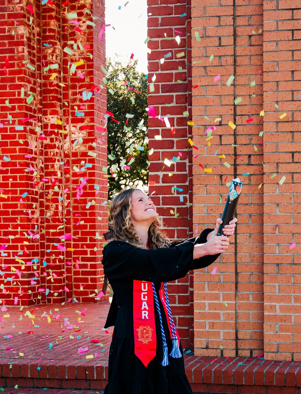 CSU Graduate tosses confetti in the air while celebrating against a red brick wall wearing a graduation stole and black attire.