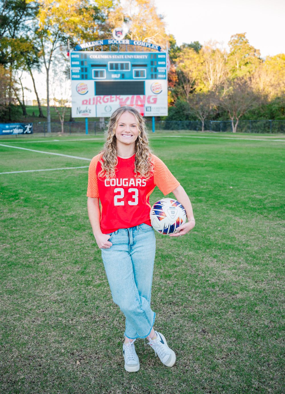 A soccer player poses with a ball and cleats in front of a scoreboard at Walden Soccer Complex at Columbus State University. College senior.