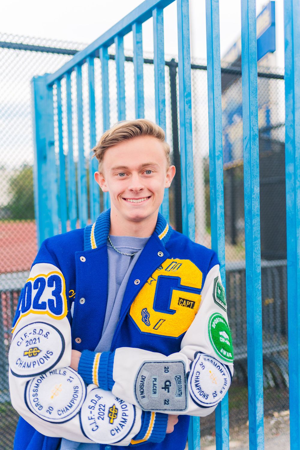 Student in blue varsity letterman jacket with patches and awards stands smiling by blue railing outdoors.