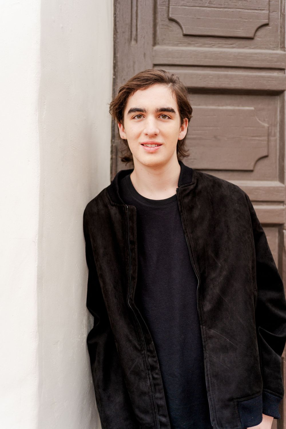 A person wearing a black jacket poses against a light-colored wall near an architectural panel.