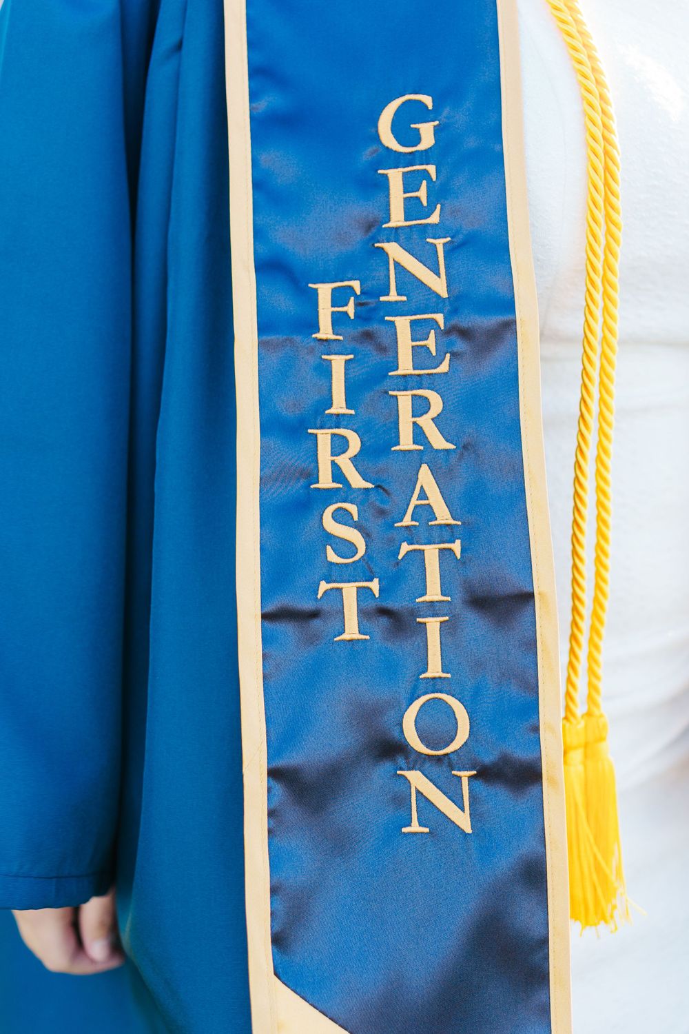 A blue graduation stole with gold lettering reading 'First Generation' and a yellow tassel visible along the edge.