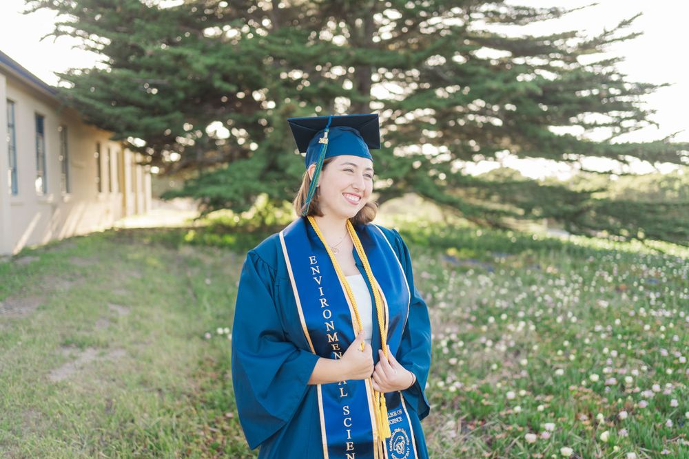 Graduate in blue cap and gown stands smiling outdoors with trees and wildflowers in background.