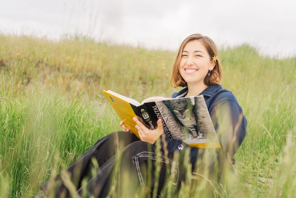 Someone reading a yellow book while sitting in a tall grass field on a sunny day, wearing a navy blue jacket.