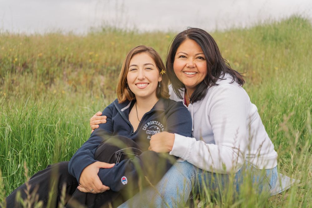 Two people sit together in a grassy field with a black dog on a sunny day outdoors.