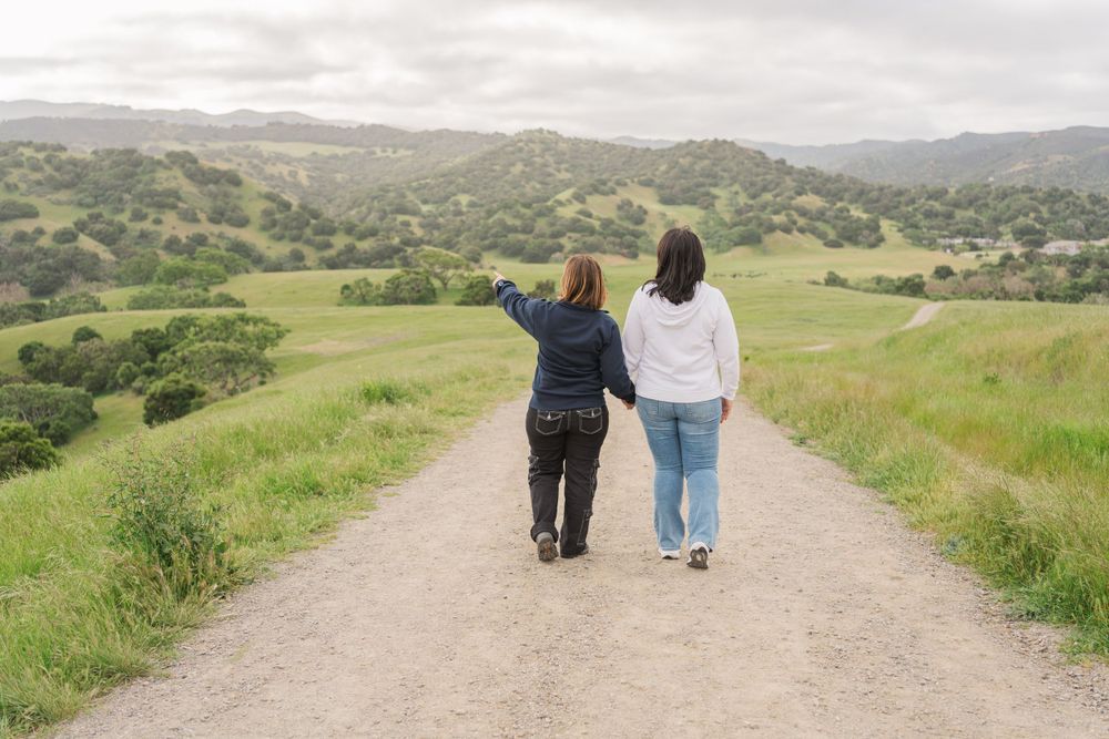 Two people walk together on a dirt trail through rolling green hills on an overcast day.