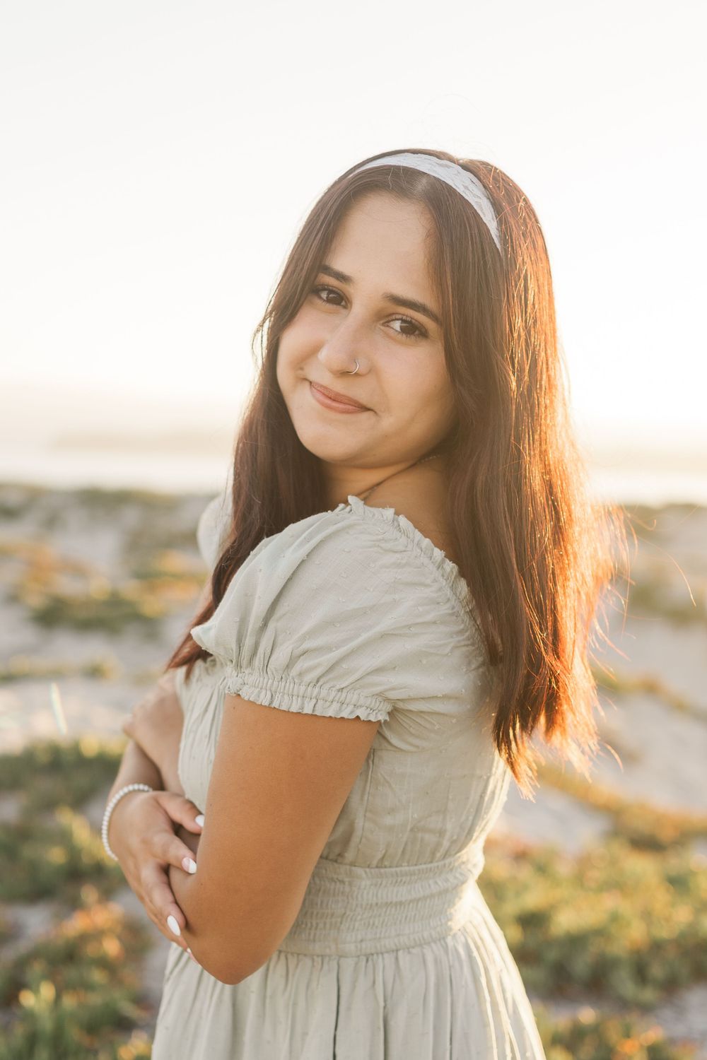 Young woman in white summer dress and hairband poses outdoors during golden hour sunset.