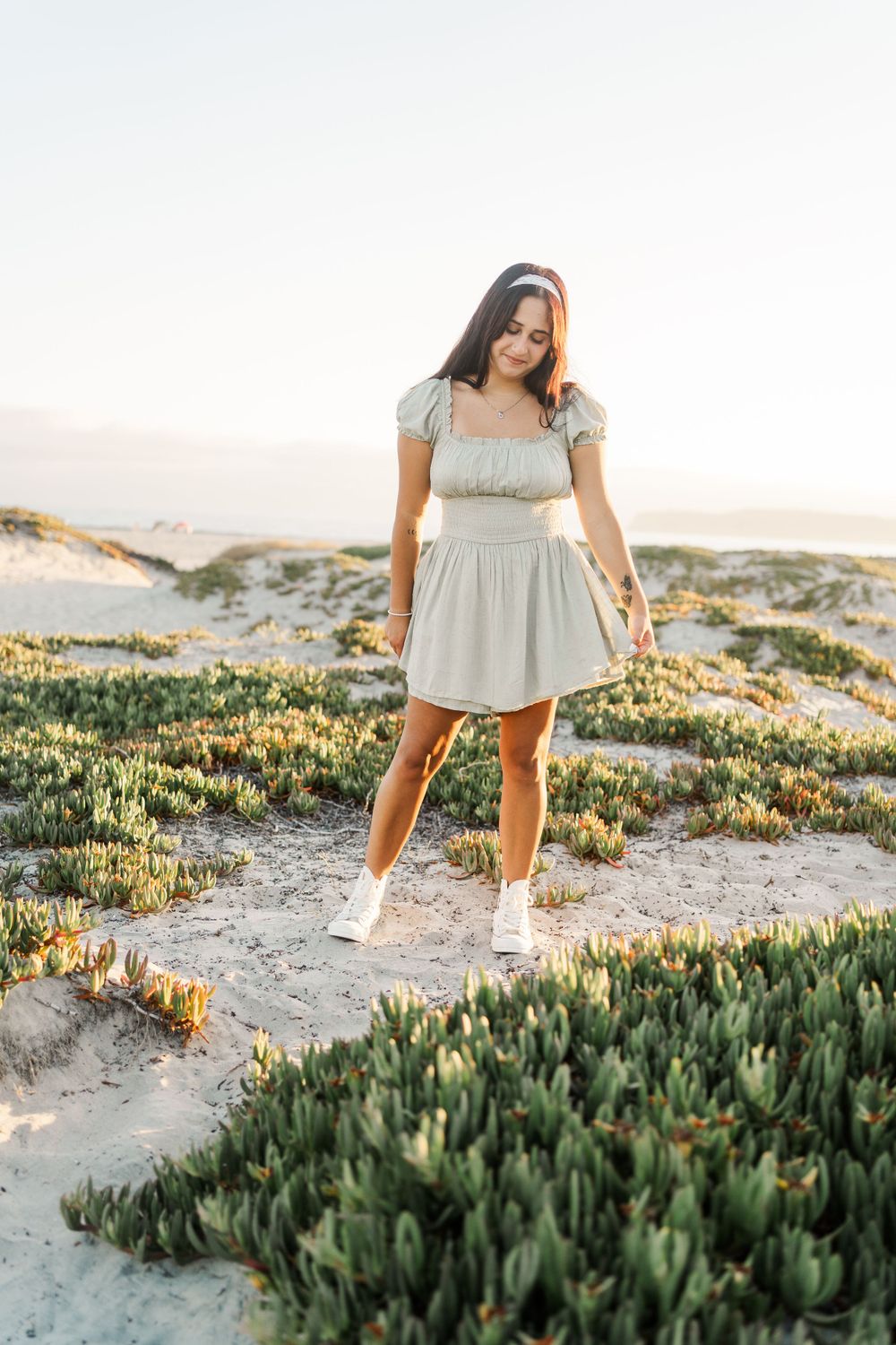 A person in a light-colored dress and white sneakers poses surrounded by coastal succulent plants at sunset.