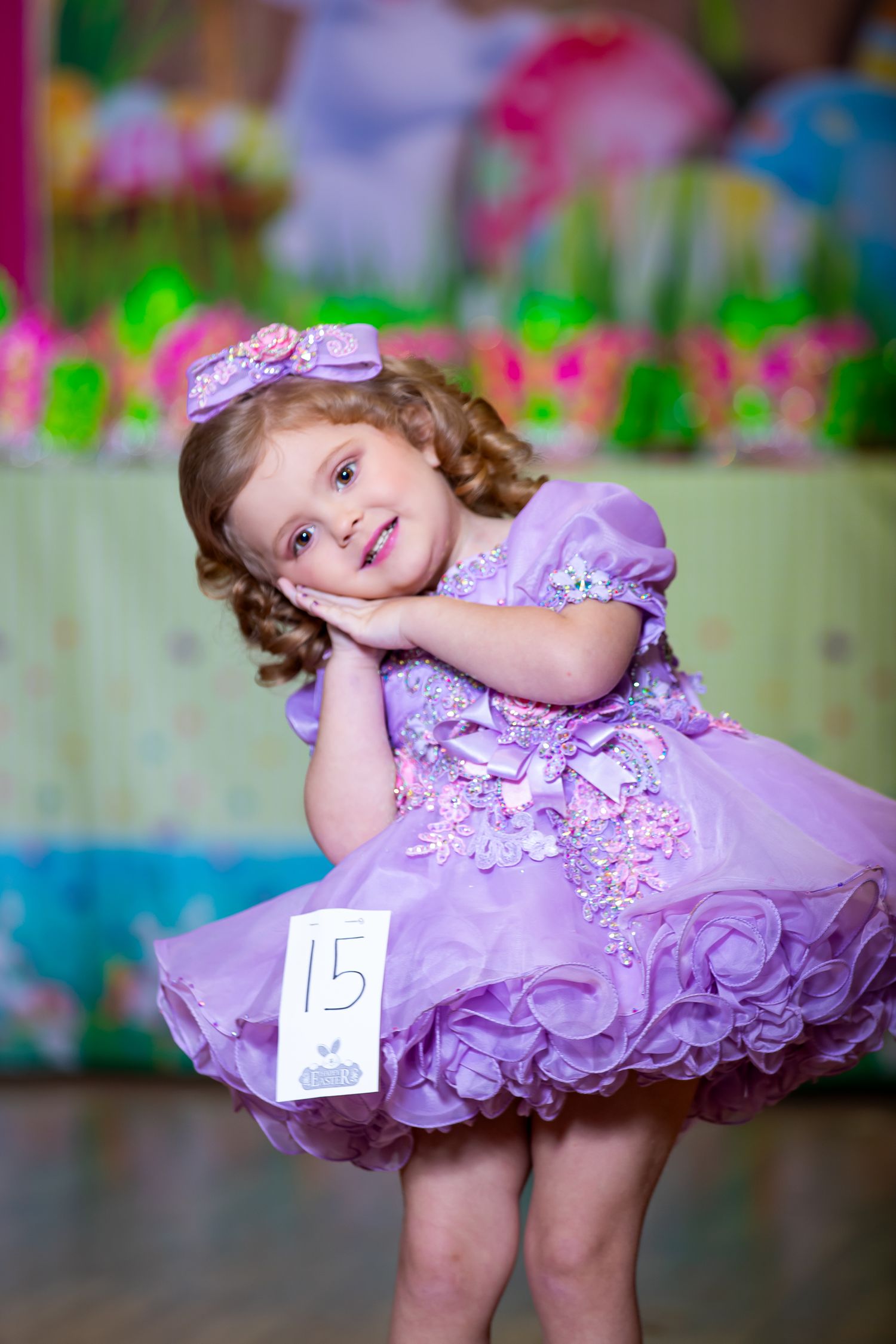 Young performer in purple pageant dress poses playfully on stage.