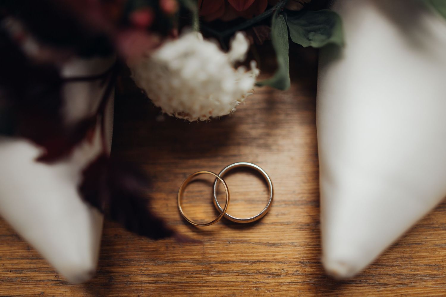 Two gold wedding bands resting on wooden surface beside a white bridal bouquet with flowers.