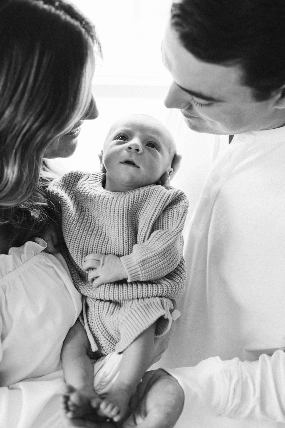 Parents share intimate moment with baby wearing knit sweater in black and white portrait.
