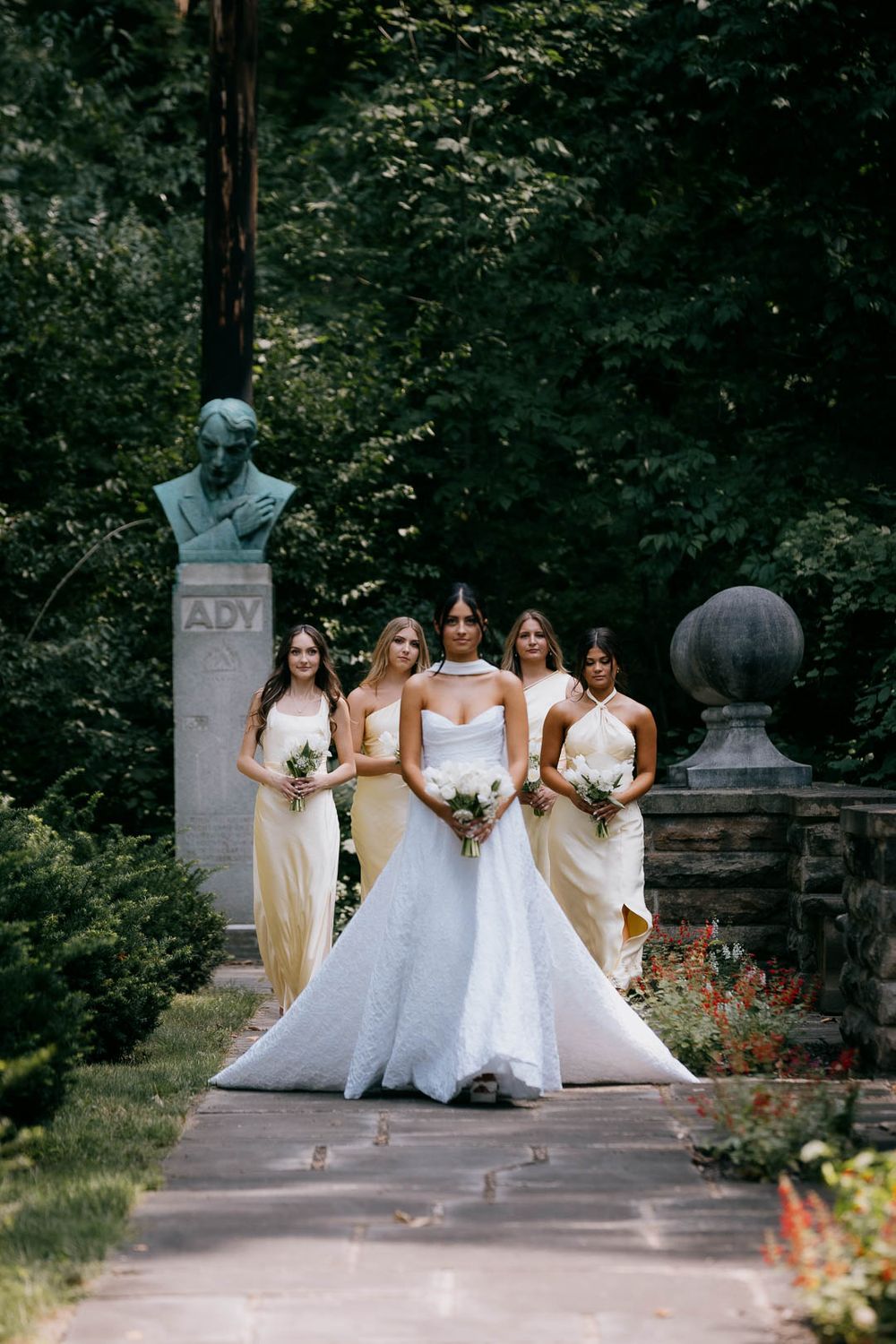Bride leads her bridesmaids through a lush garden at Ariel Pearl Center during wedding portrait, captured by Cleveland wedding photographer.