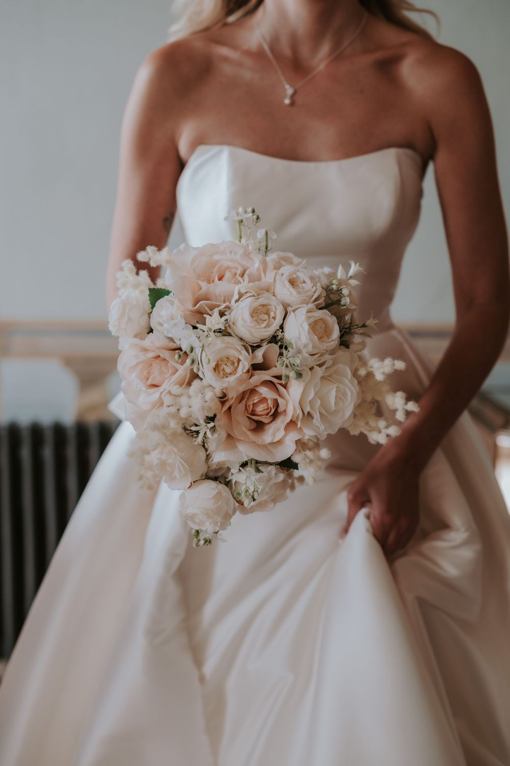 A romantic bridal bouquet of blush and white roses held against an elegant white strapless wedding gown.