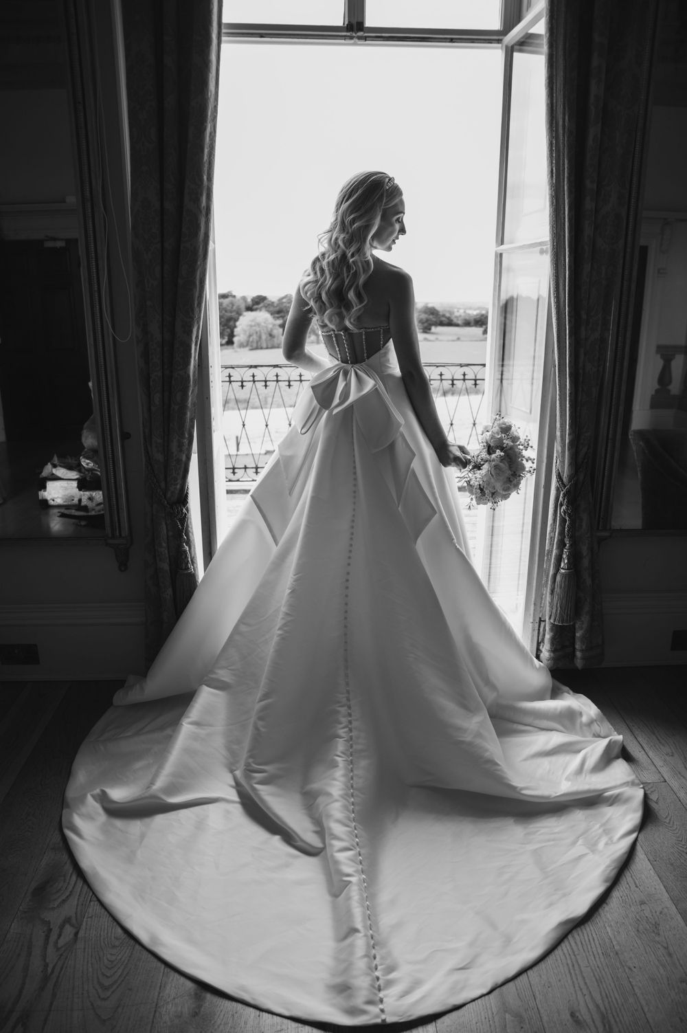 A black and white elegant bridal shot shows a flowing wedding gown with long train silhouetted against a bright window.