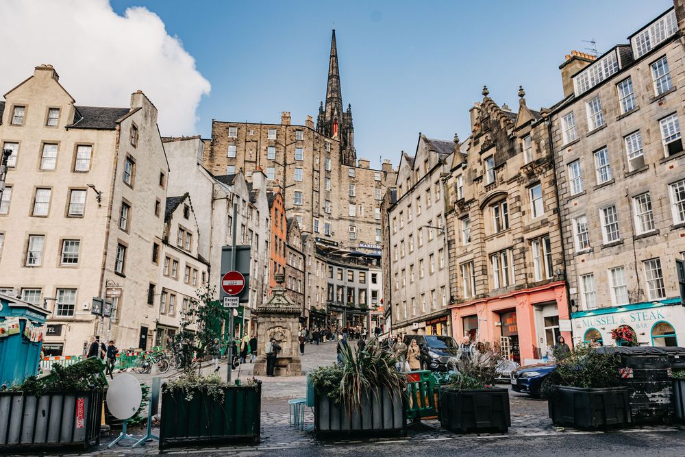 Charming cobblestone square in Edinburgh's Old Town with historic stone buildings, lush greenery, and a Gothic spire in the background.
