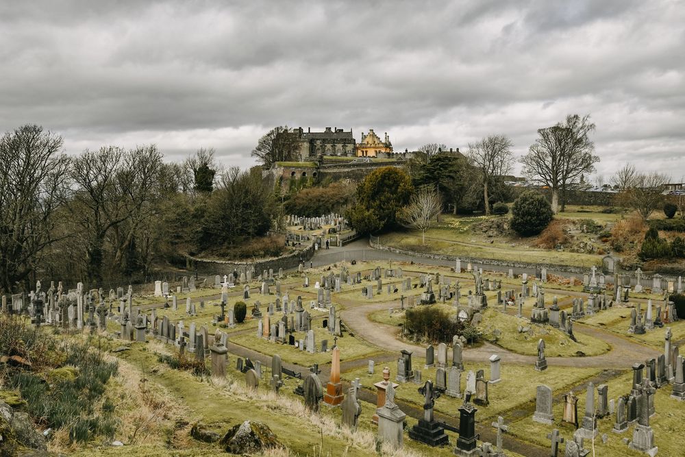 A historic castle atop a hill overlooking a large cemetery with gravestones under cloudy skies.