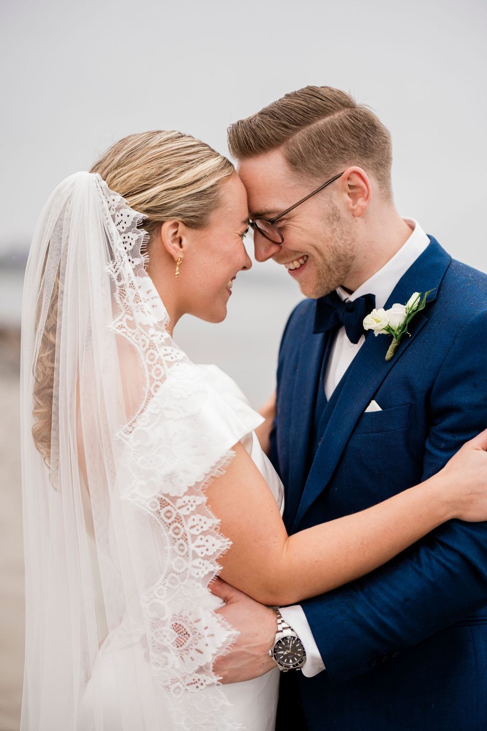 Smiling bride and groom in navy suit touch foreheads tenderly outdoors on their wedding day.
