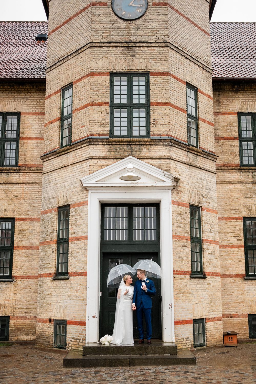 Bride and groom holding clear umbrellas stand at the entrance of a historic brick building on their wedding day.