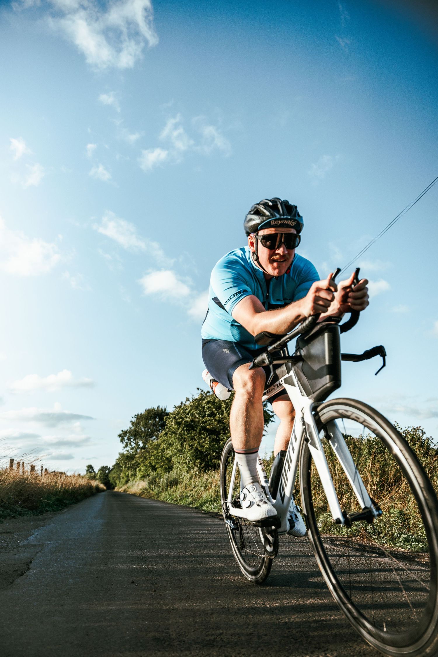A cyclist in blue jersey rides a white road bike along a scenic uphill road on a sunny day.