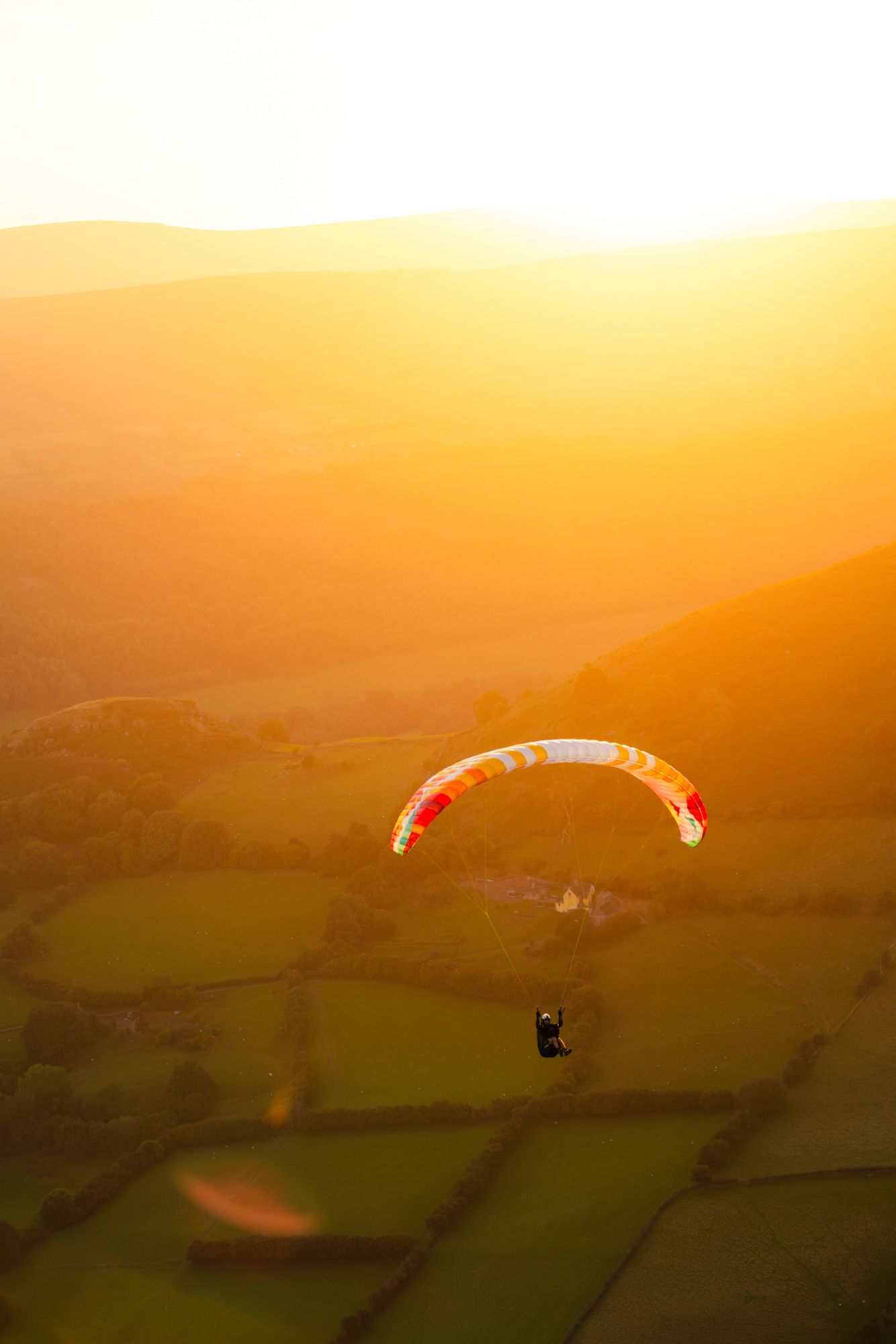 Paraglider with red and white canopy gliding over golden landscape at sunset.