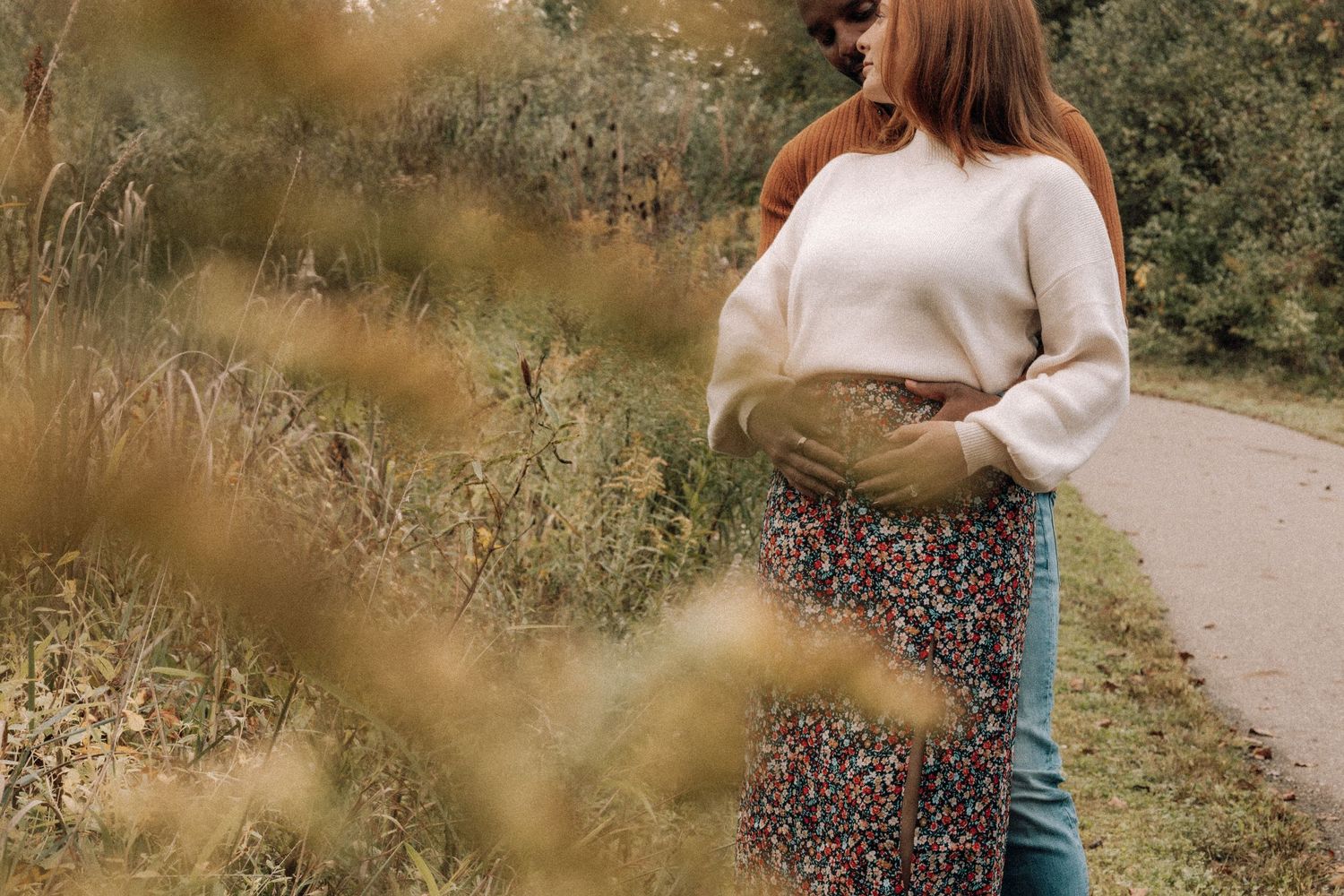 A pregnant woman in a cream sweater and floral skirt cradles her baby bump on an outdoor path.