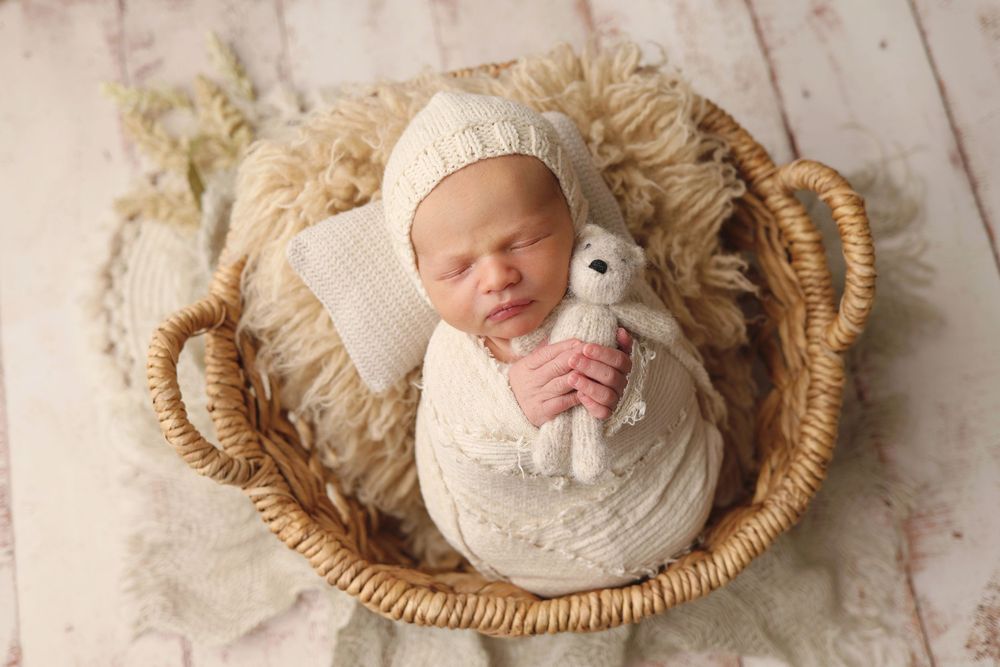 Swaddled newborn baby sleeping in wicker basket holding a knit teddy bear.