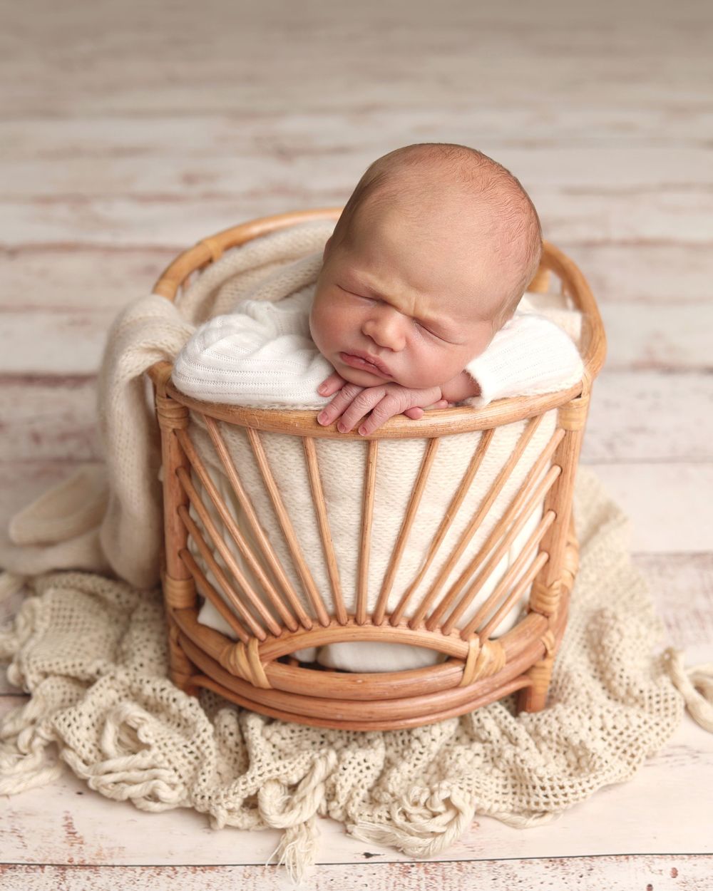 Sleeping newborn baby resting chin on hands in a rattan basket with cream wrap.