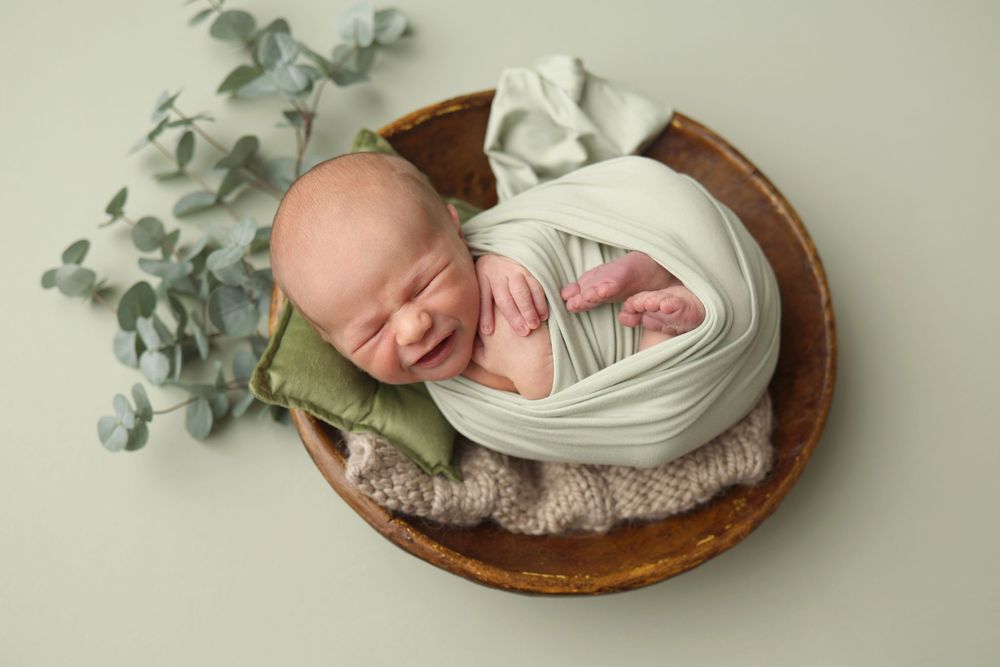 Smiling newborn baby wrapped in white swaddle sleeping in wooden bowl with green eucalyptus leaves.