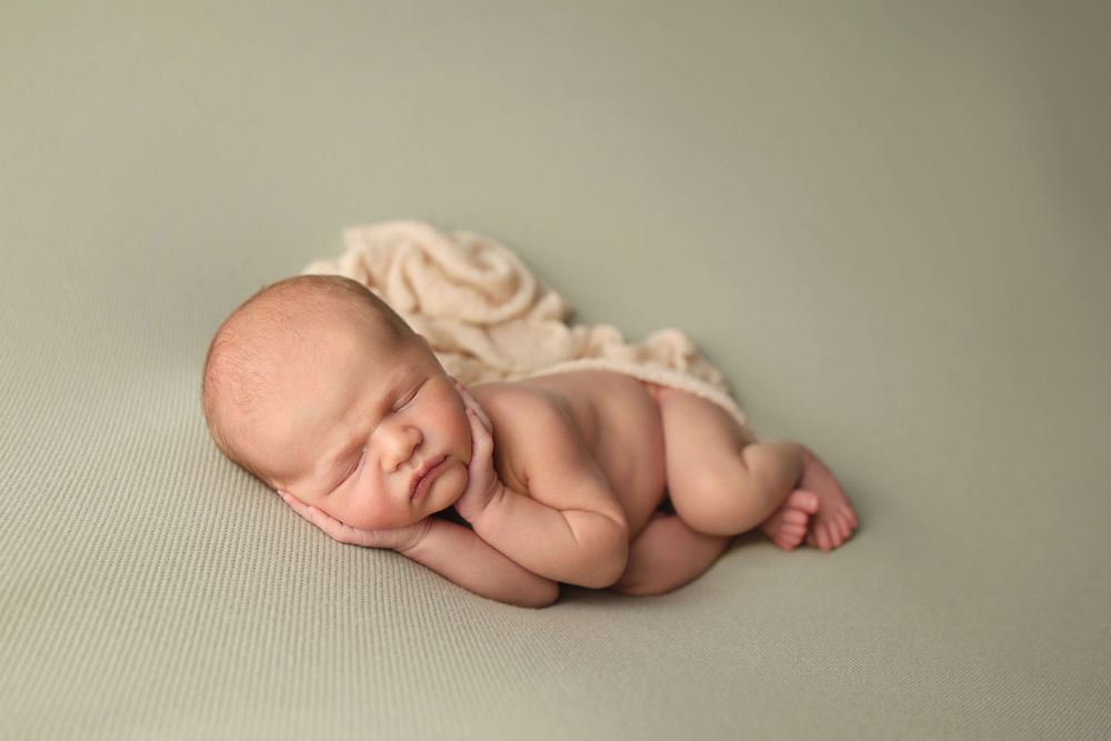 Smiling newborn baby curled up sleeping on a sage green textured backdrop with cream wrap.
