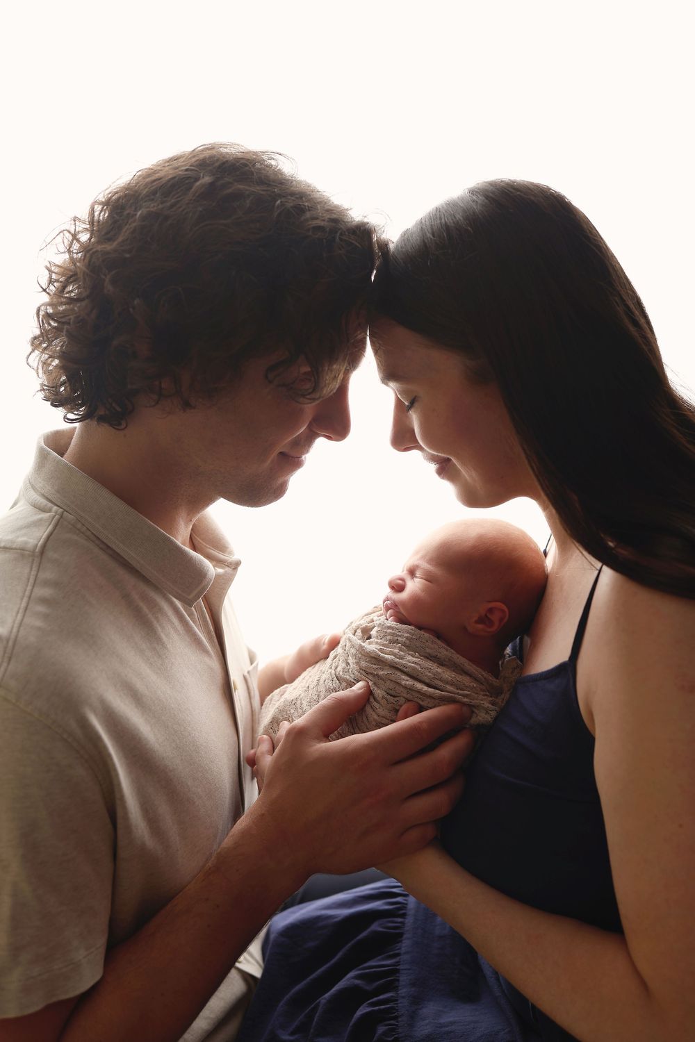 Silhouetted parents touching foreheads together while cradling their swaddled newborn baby in light.