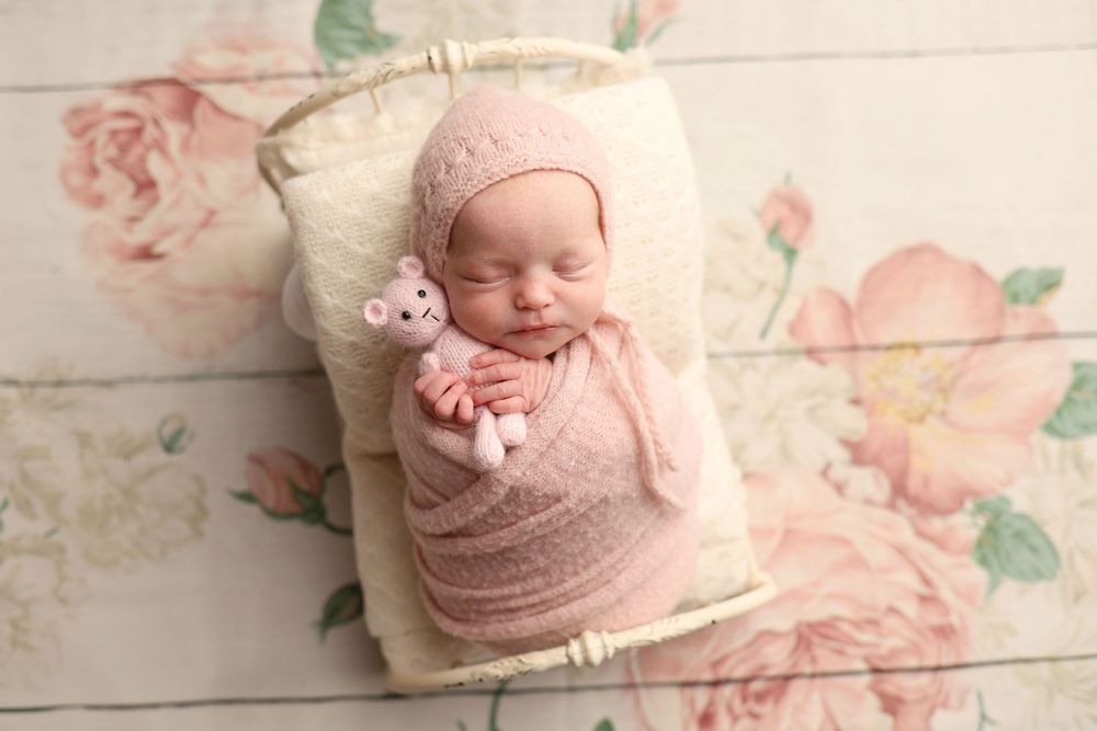 Sleeping newborn baby wrapped in pink knit swaddle wearing bonnet, holding tiny stuffed animal in a small wooden basket.