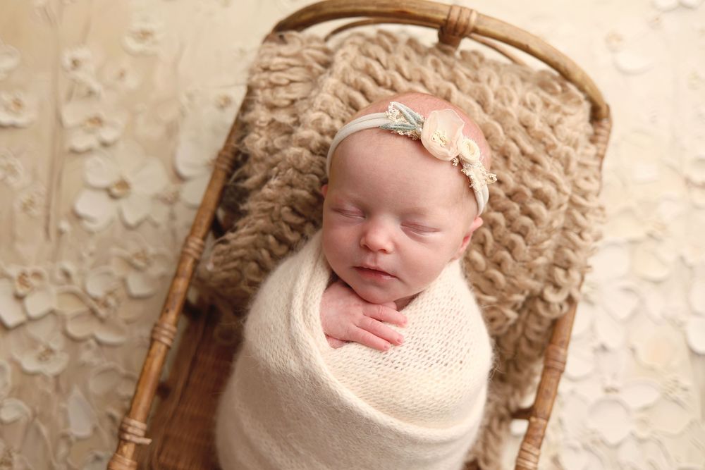 Newborn baby swaddled in cream wrap, sleeping peacefully in a wicker basket with a floral headband.