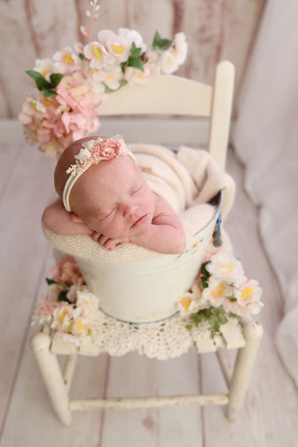 Sleeping newborn baby girl in cream bucket on white chair surrounded by pink and white flowers wearing floral headband.